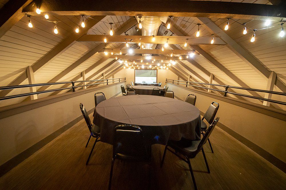 Rooftop event space with round tables, chairs, and string lights. Wooden beams, rope railings, and a projector screen.