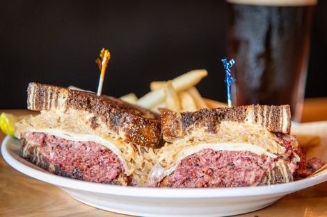 Reuben sandwich on a plate with fries, toothpick, and dark beer in a glass.