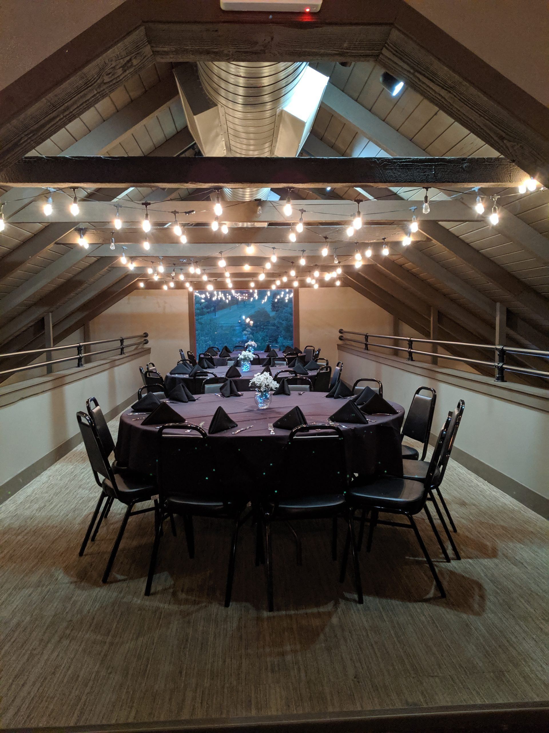 Event space with round table set for a gathering, dark linens, black chairs, and string lights.