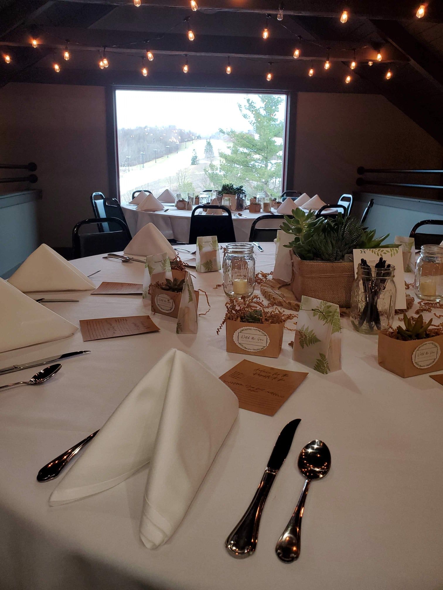 Wedding reception table setting with white linens, napkins, and centerpieces near a window.