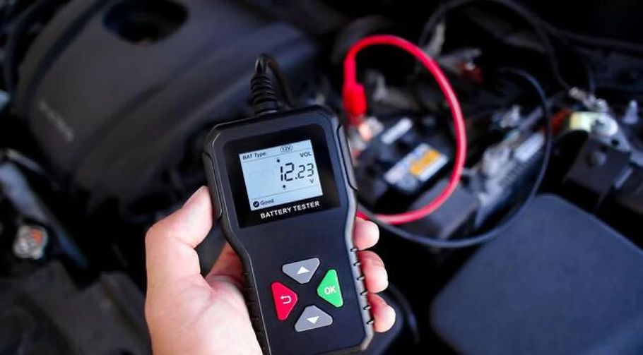 Hand holding a car battery tester near engine wires and a red cable