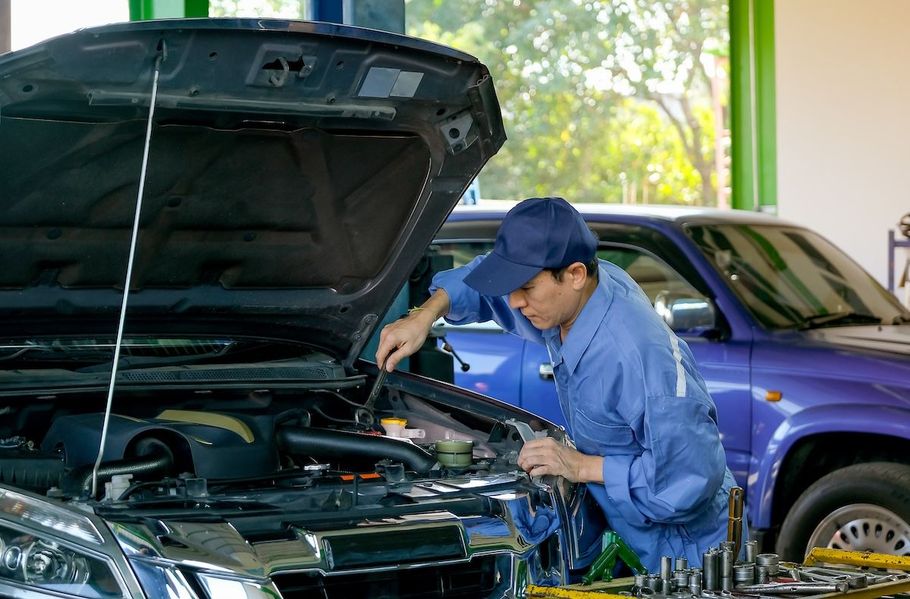 Mechanic in blue coveralls inspecting a car engine with hood open beside a blue SUV in a garage