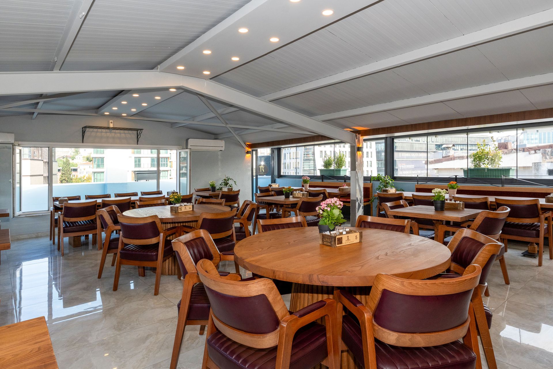 Mays Otel | Restaurant dining area with round wooden tables, cushioned chairs, and retractable roof.