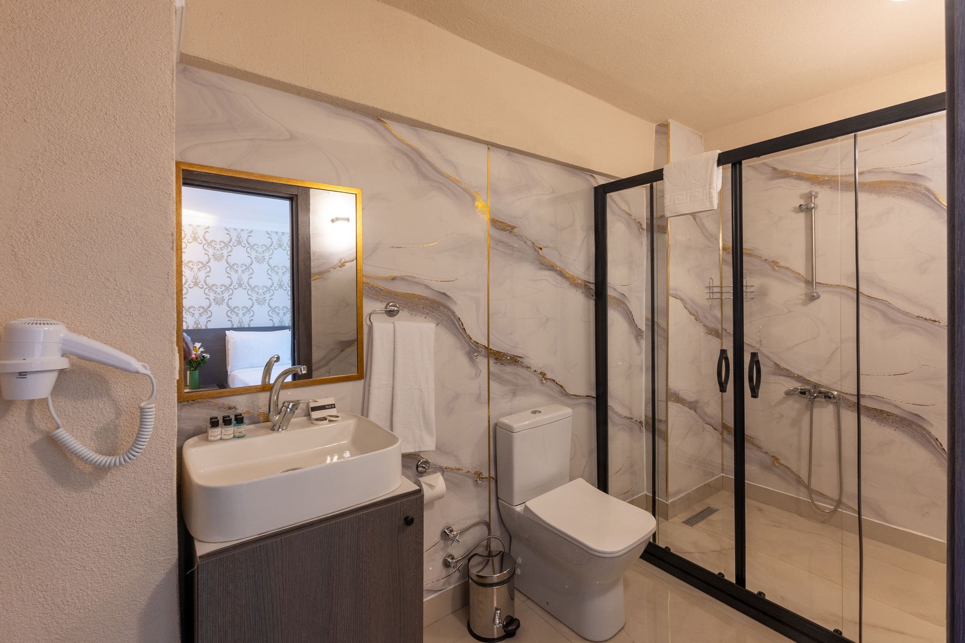 Mays Otel | Bathroom with marble-effect wall, glass shower, toilet, sink with mirror, and hairdryer.
