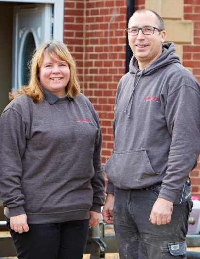 Two people in gray hoodies standing outside a brick building, smiling at the camera.
