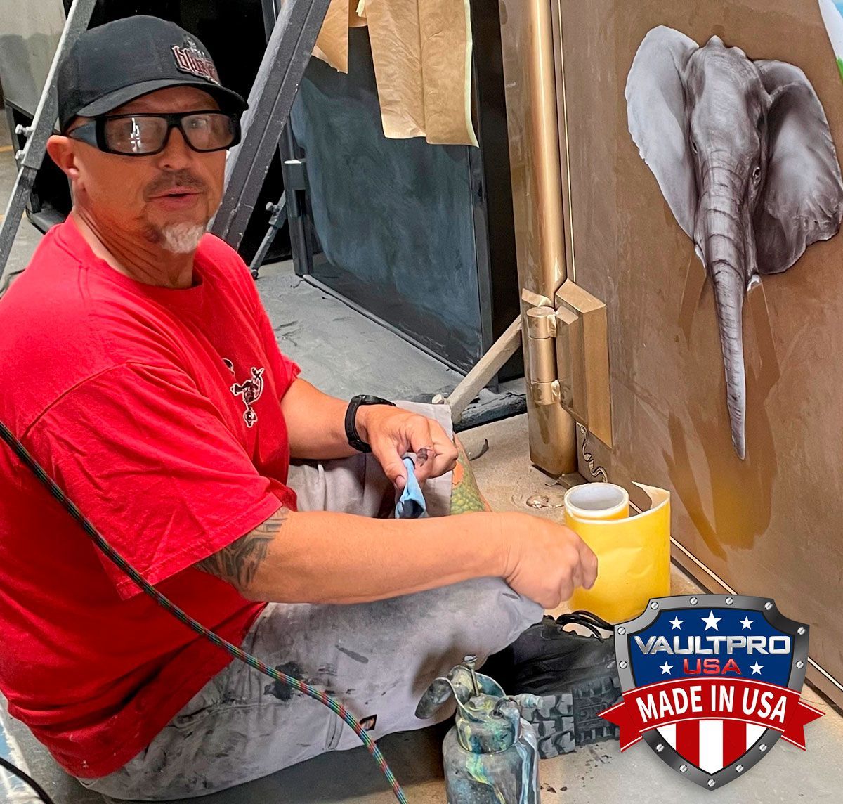 Artist painting a wall mural, seated with tools and yellow paint bucket, beside a gray elephant sketch.