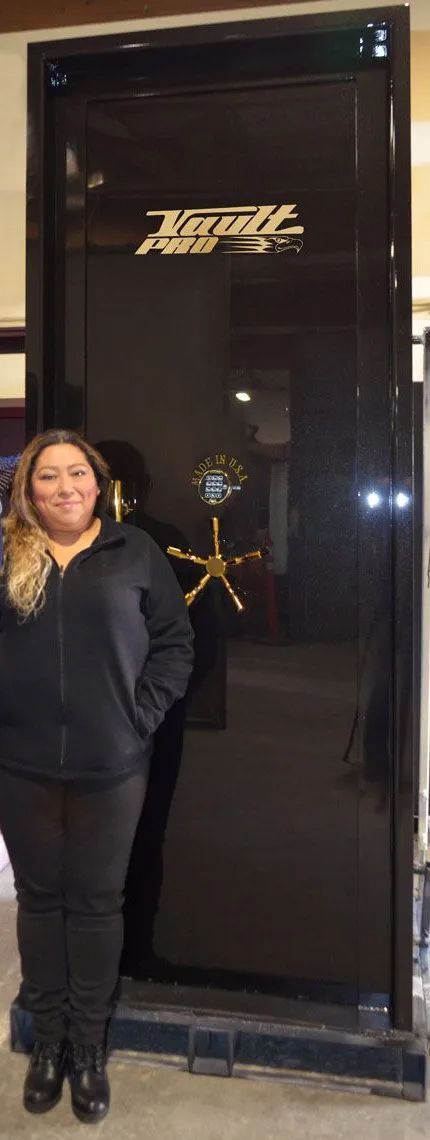 Two people posing indoors beside a black glass door with a gold logo, one in blue coveralls and one in black.
