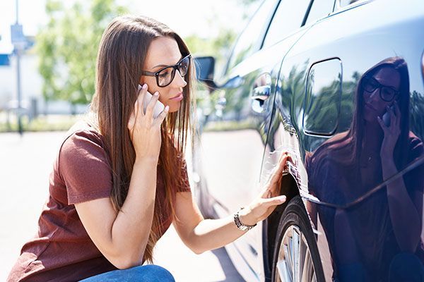 Woman in glasses on phone, examining dent in car door outdoors. | Auto Clinic Care