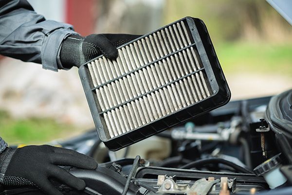 Gloved hands holding a dirty car air filter near an open car engine. | Auto Clinic Care