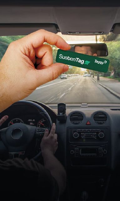 Mão segurando um chaveiro verde dentro de um carro, com a estrada visível através do para-brisa.