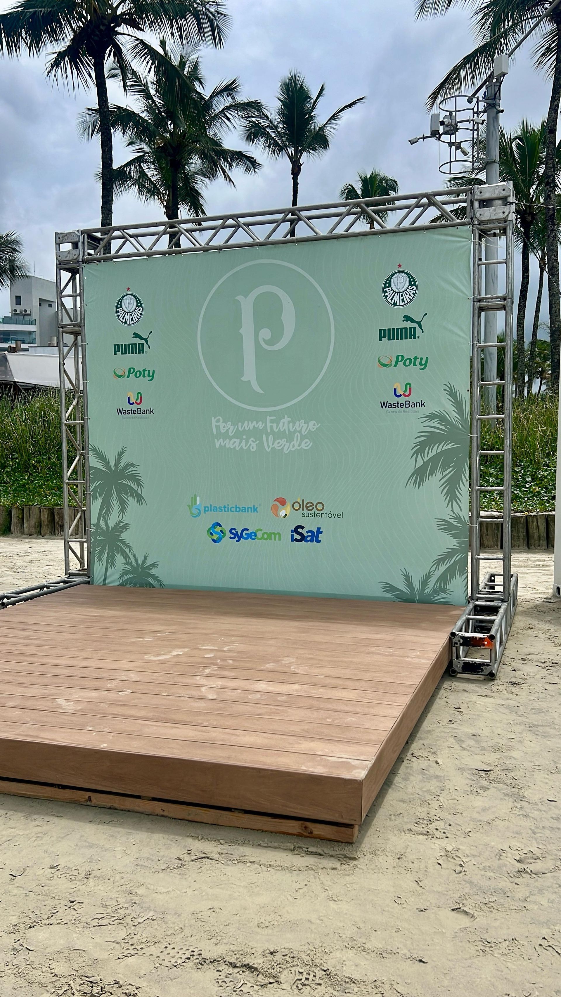 Palco para eventos ao ar livre com um painel de fundo verde claro com a marca e plataforma de madeira na areia, emoldurado por palmeiras.