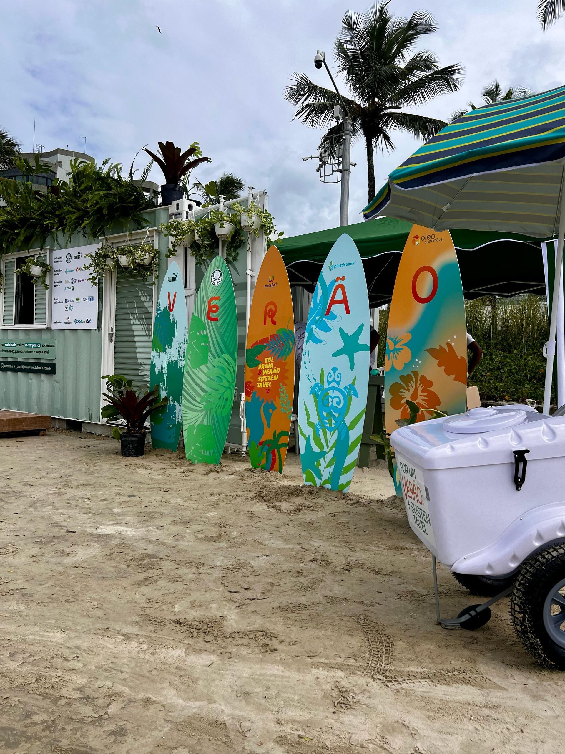 Pranchas de surf coloridas com letras estão ao lado de uma cabana de praia tropical e uma scooter branca sob palmeiras.