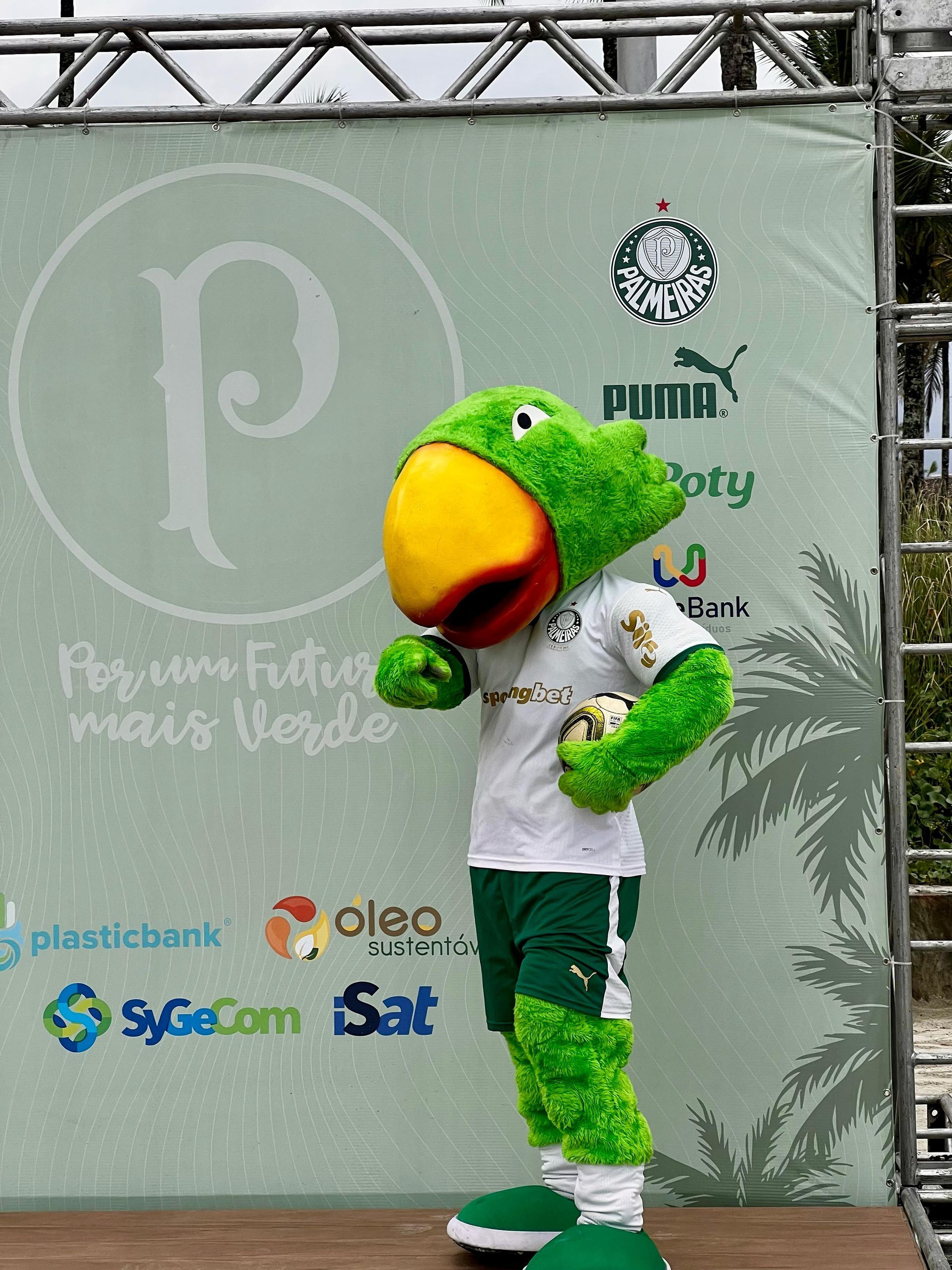 Mascote de papagaio verde com camisa branca em um palco, com um grande bico laranja e logotipos de patrocinadores ao fundo.