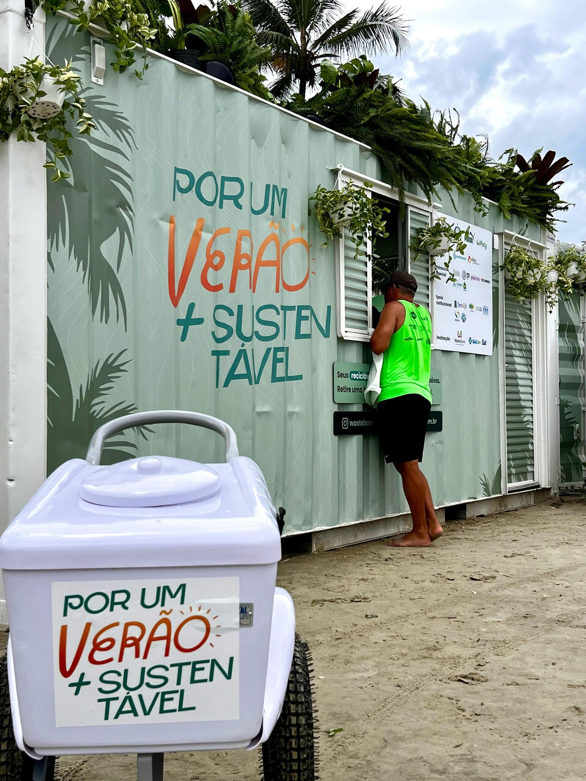 Person in neon green shirt standing by a white kiosk with “POR UM VERÃO + SUSTENTÁVEL” text.
