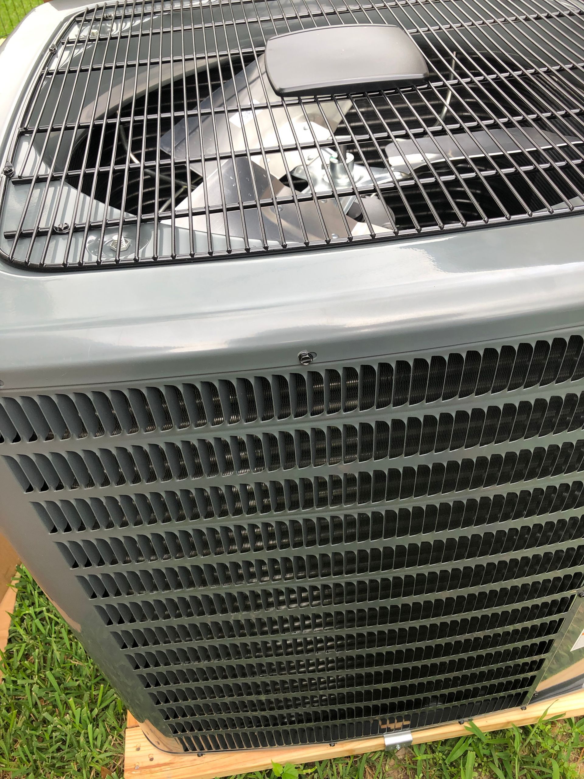 Close-up of a gray air conditioner unit with black grille and fan visible on top, outdoors on grass