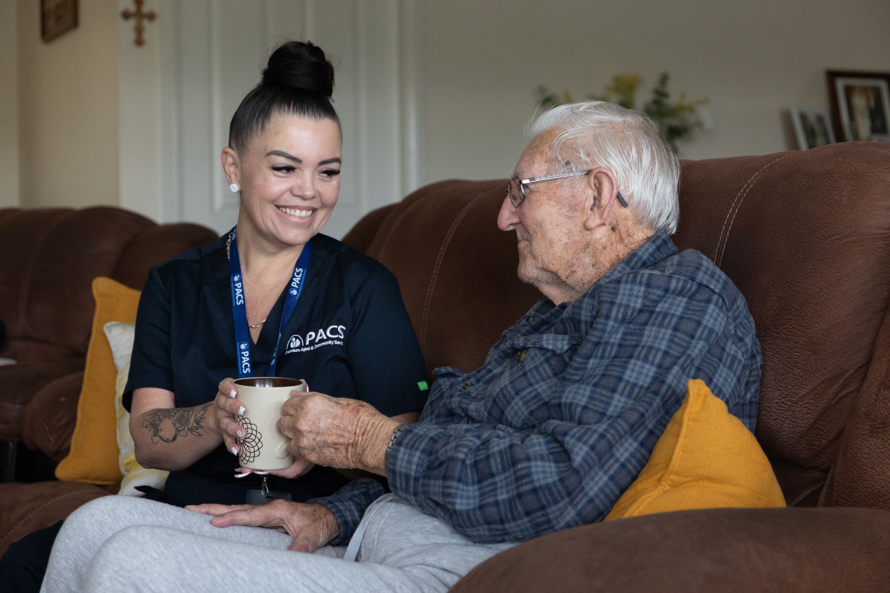 Caregiver Offering a Mug to an Elderly Person on a Sofa; Both Smiling — Premium Aged & Community Services In Tamworth, NSW