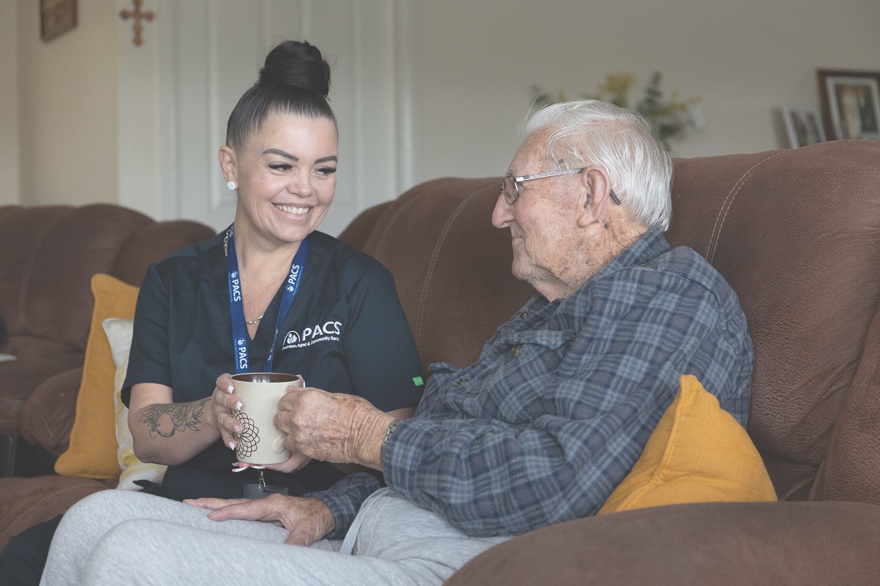Woman in a Black Uniform Smiles at an Elderly Man as She Offers Him a Mug — Premium Aged & Community Services In Gosford, NSW