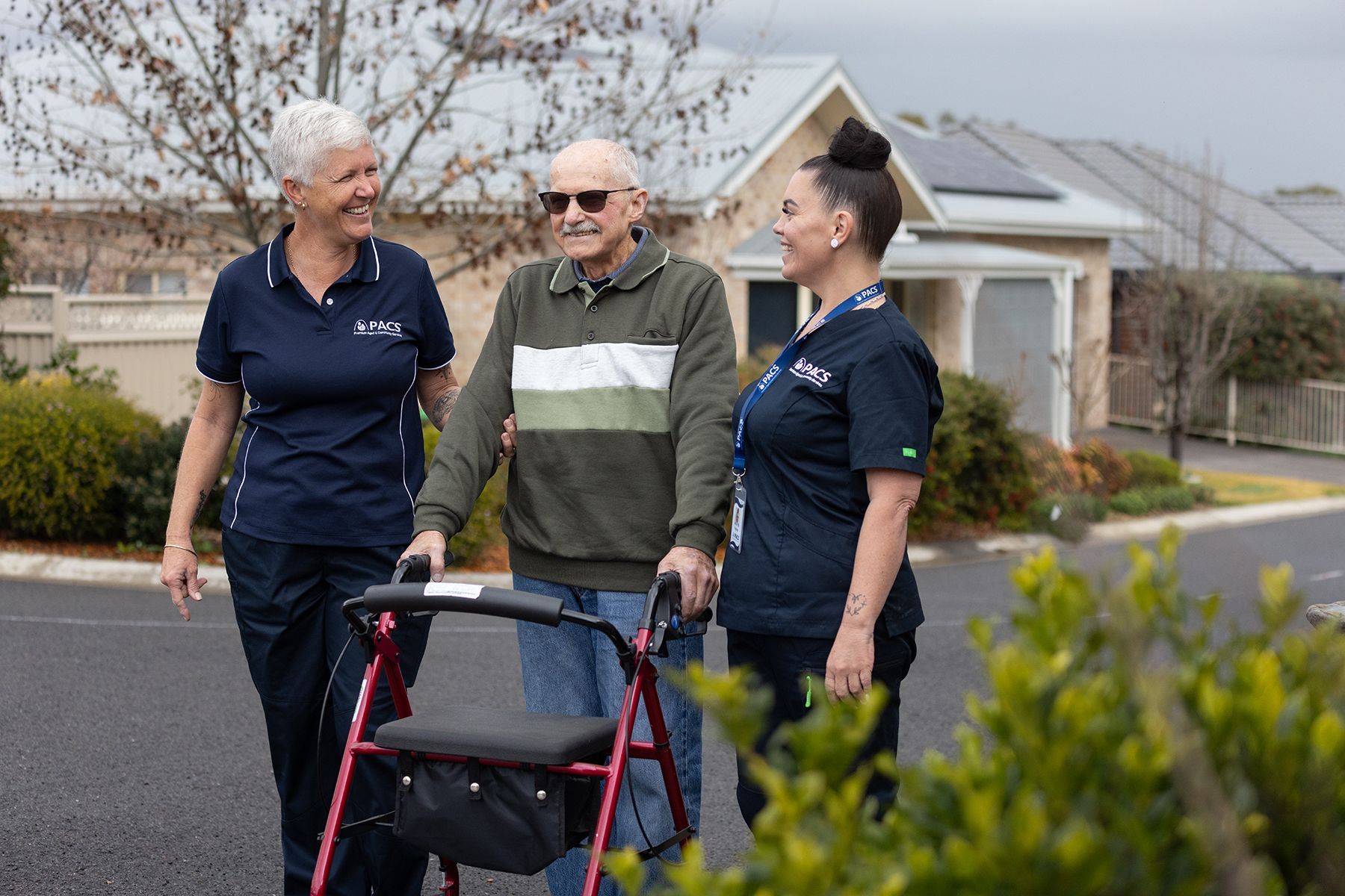 Two Caregivers Assist an Older Person Using a Walker Outdoors, Smiling — Premium Aged & Community Services In Quirindi, NSW