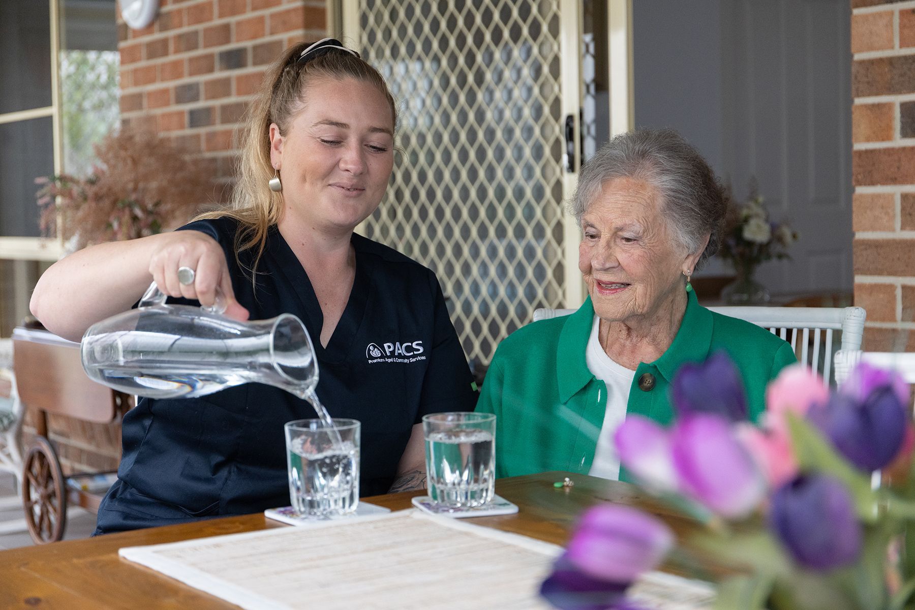 Caregiver Pouring Water for an Elderly Woman Sitting Outdoors at a Table — Premium Aged & Community Services In Tamworth, NSW