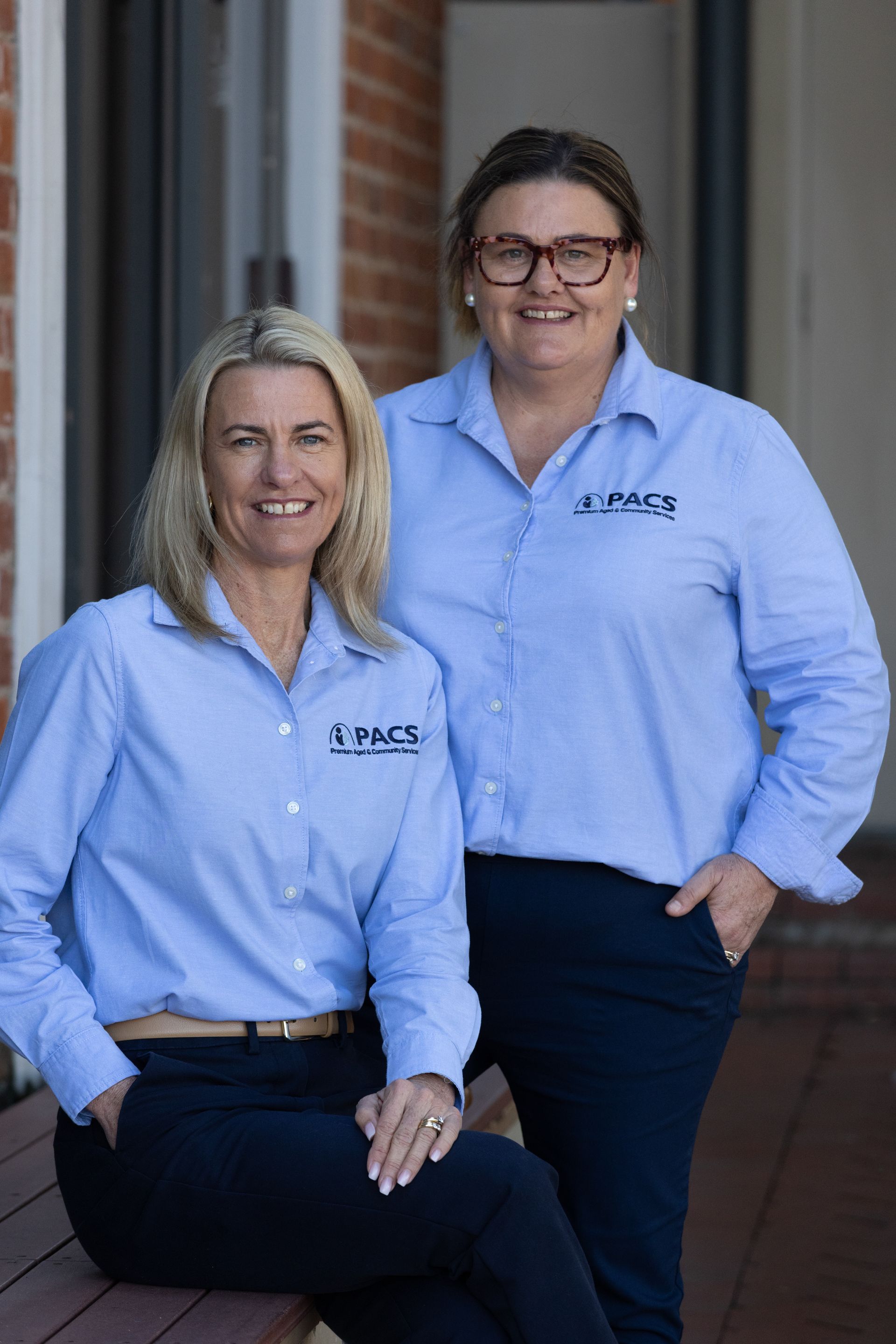 Two Women in Blue Shirts, Smiling, Posing Together Outside a Building — Premium Aged & Community Services In Tamworth, NSW