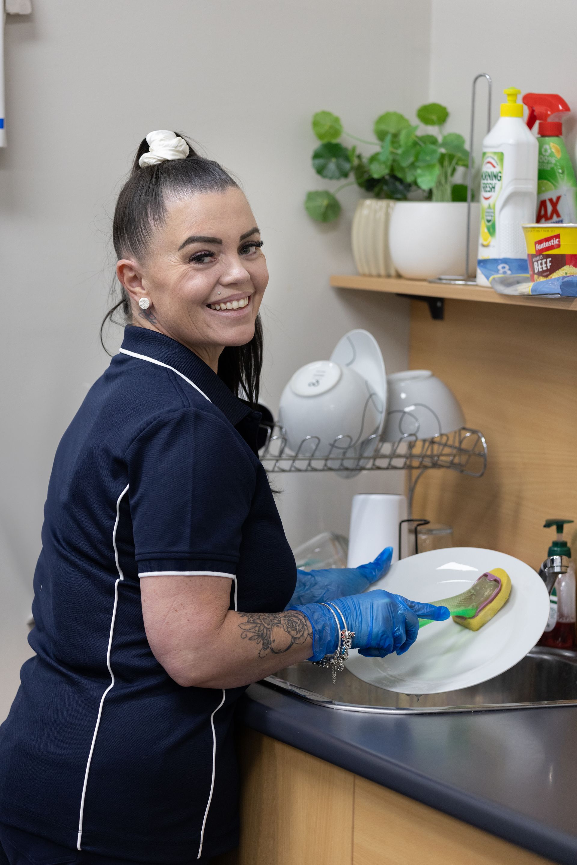 Woman in Blue Scrubs Washes Dishes, Smiling in a Kitchen — Premium Aged & Community Services In Tamworth, NSW