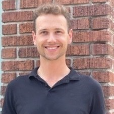 Artur in a black shirt is smiling in front of a brick wall .