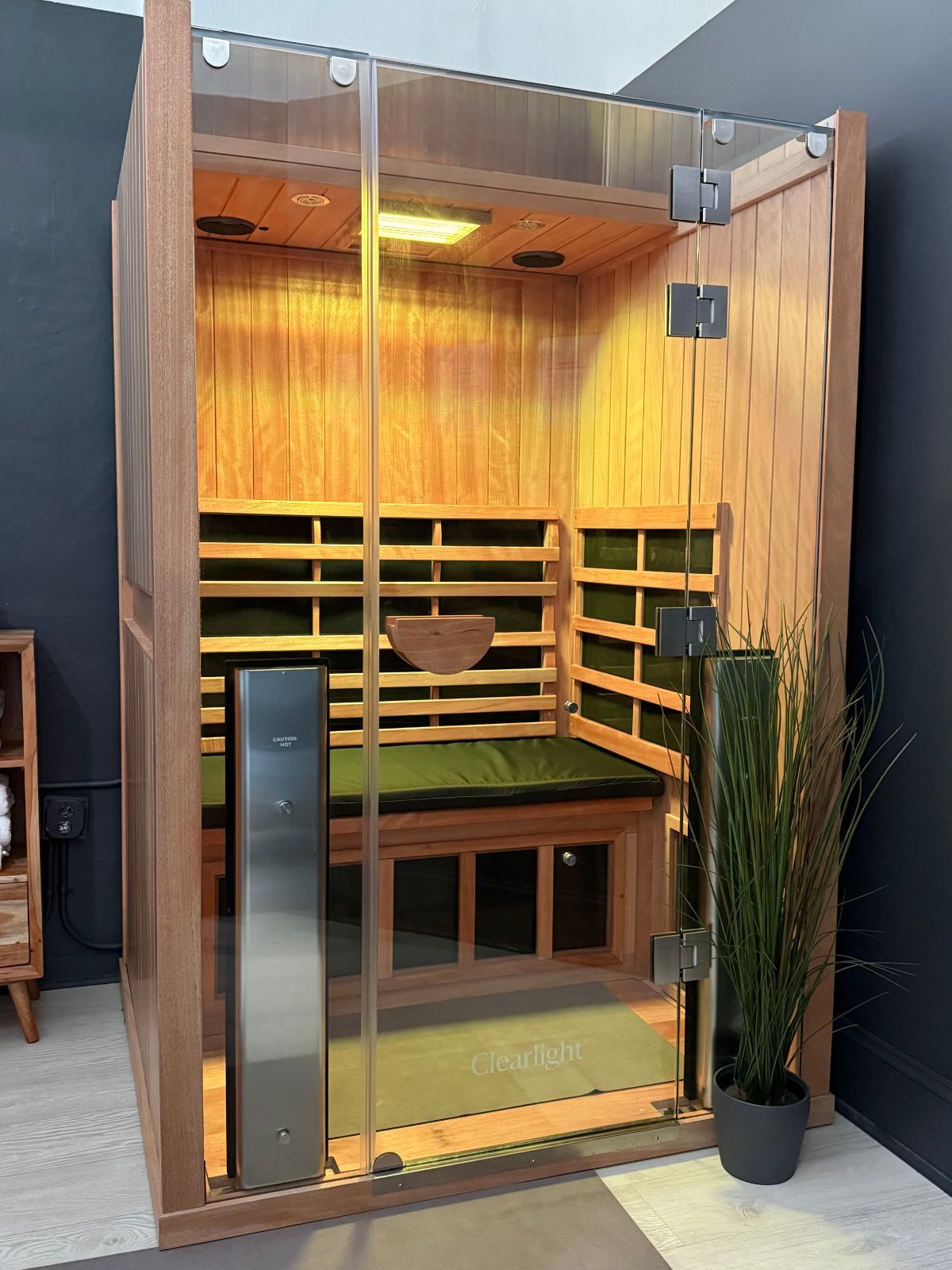 Wooden infrared sauna with glass door, bench, and control panel. Inside are dark green cushions and a wooden bowl.