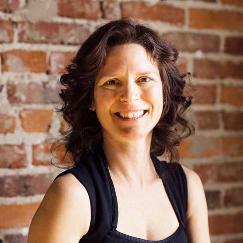 A woman in a black tank top is smiling in front of a brick wall.
