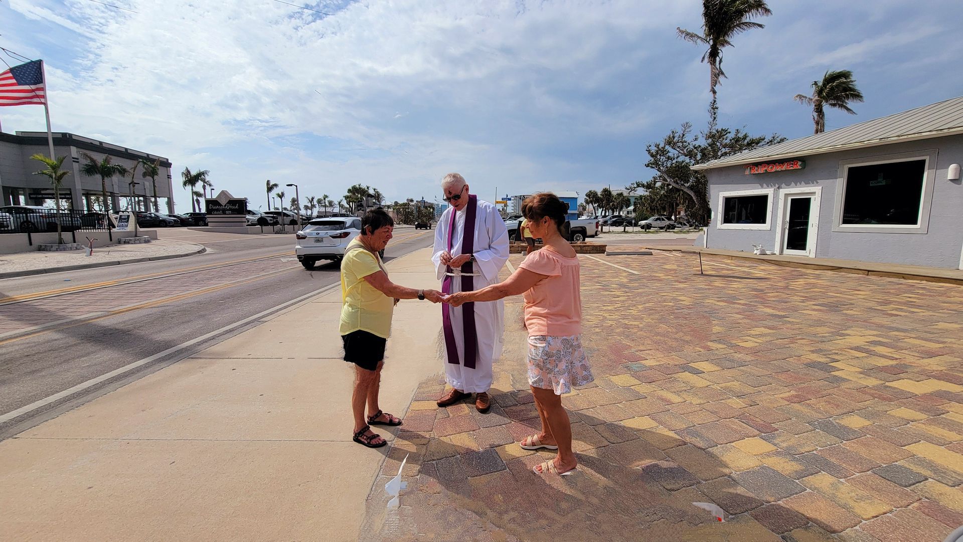 A priest blesses two girls holding hands outdoors near a road and buildings.