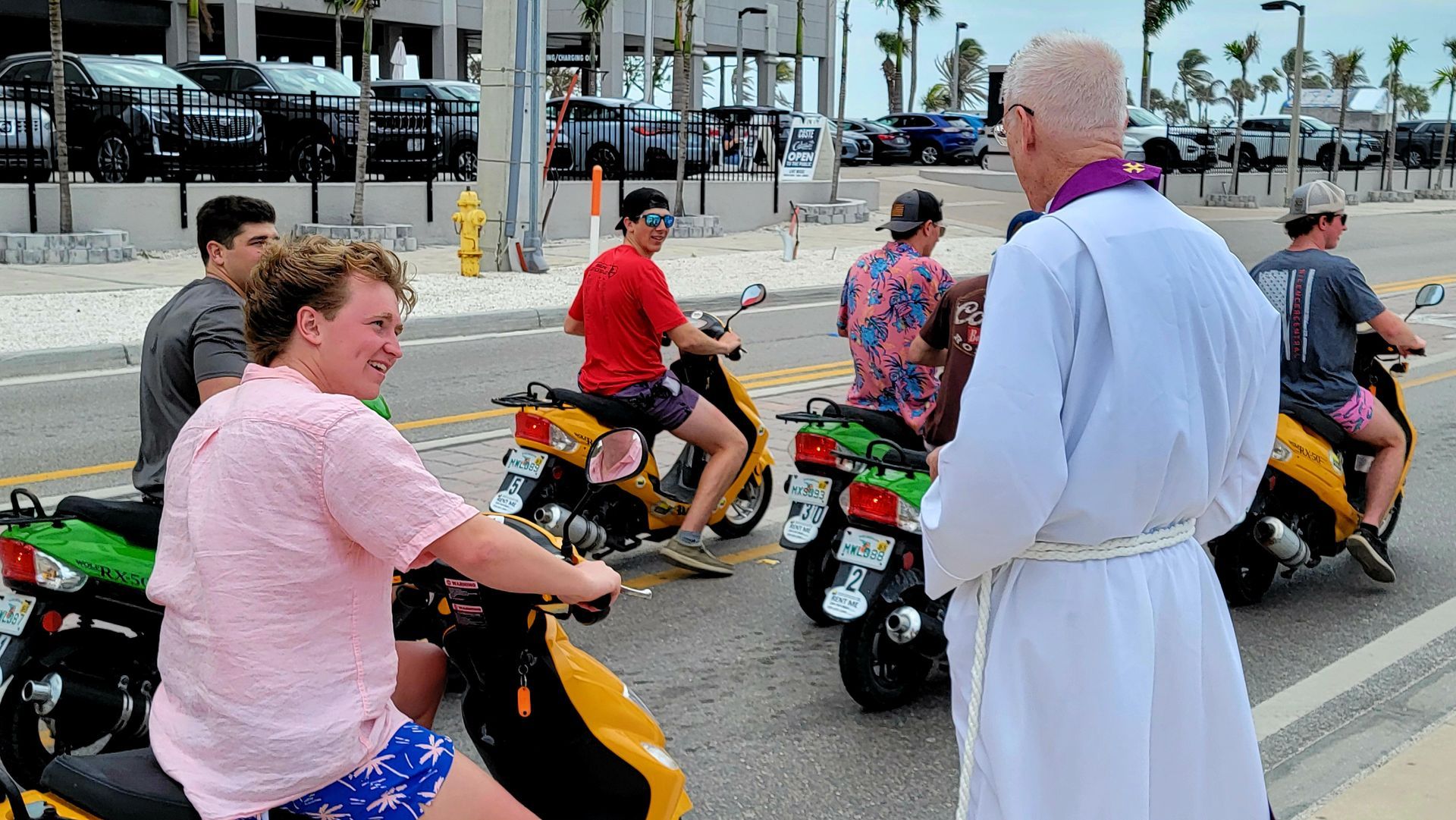 A priest in white robe talking to a group of young men on colorful scooters on a sunny street.