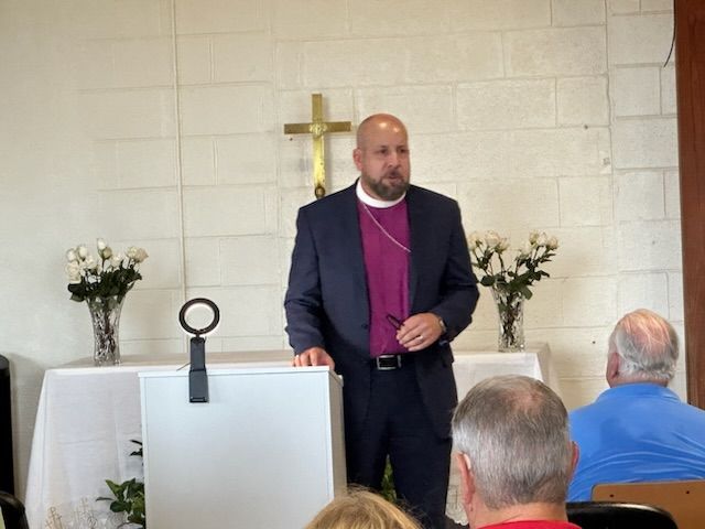 Man in suit and clerical collar speaks at podium, with a cross and flowers behind him, in a church.