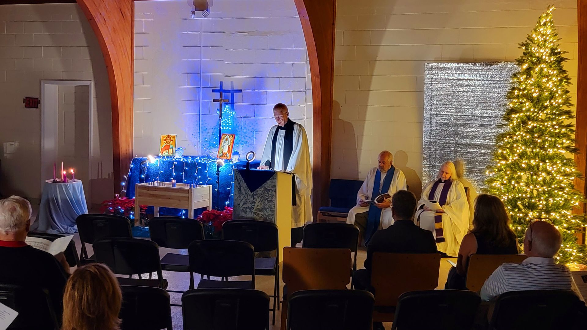Church service with pastor speaking, Christmas tree lit, altar with decorations, people seated.