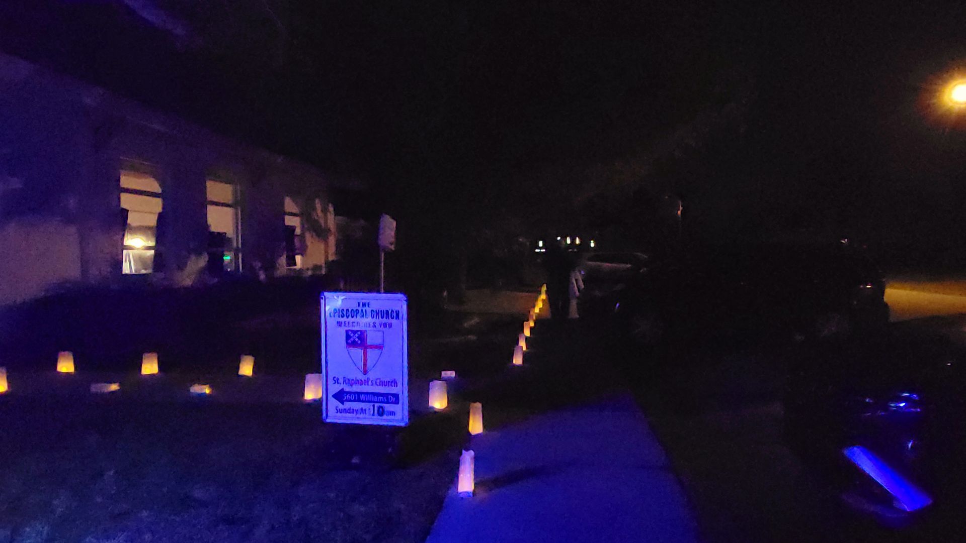 Night scene with illuminated walkway. Sign directs to event. Buildings and lights in the background.