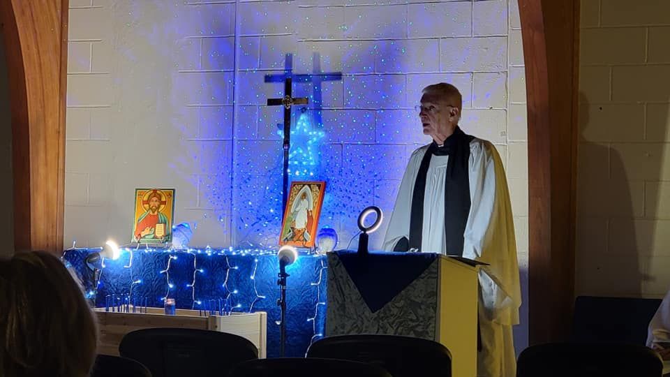 Clergyman giving a sermon at a podium in front of a cross, illuminated by blue lights and religious icons.