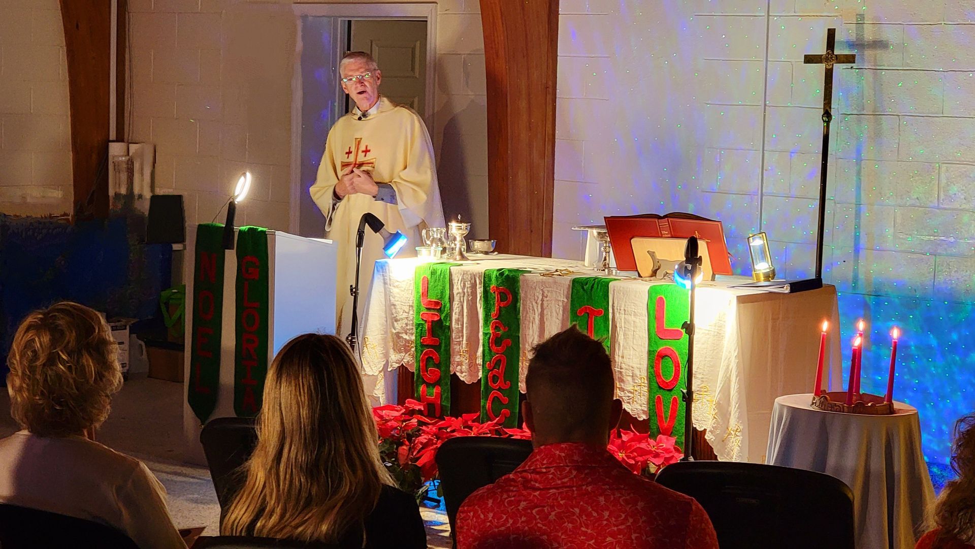 A priest speaks at a church altar with an audience. Lit candles and decorations are present.