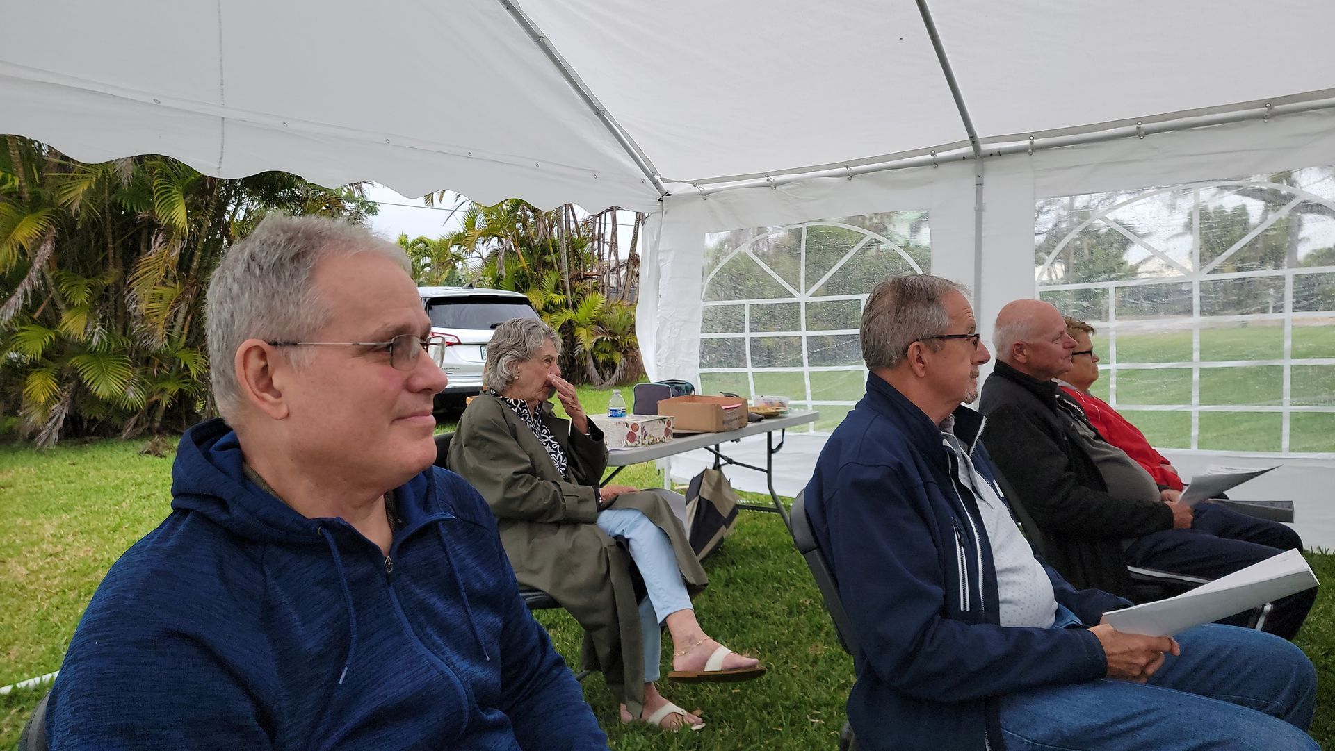 Group of people seated under a white tent, outdoors on grass. Several are looking to the right, listening.
