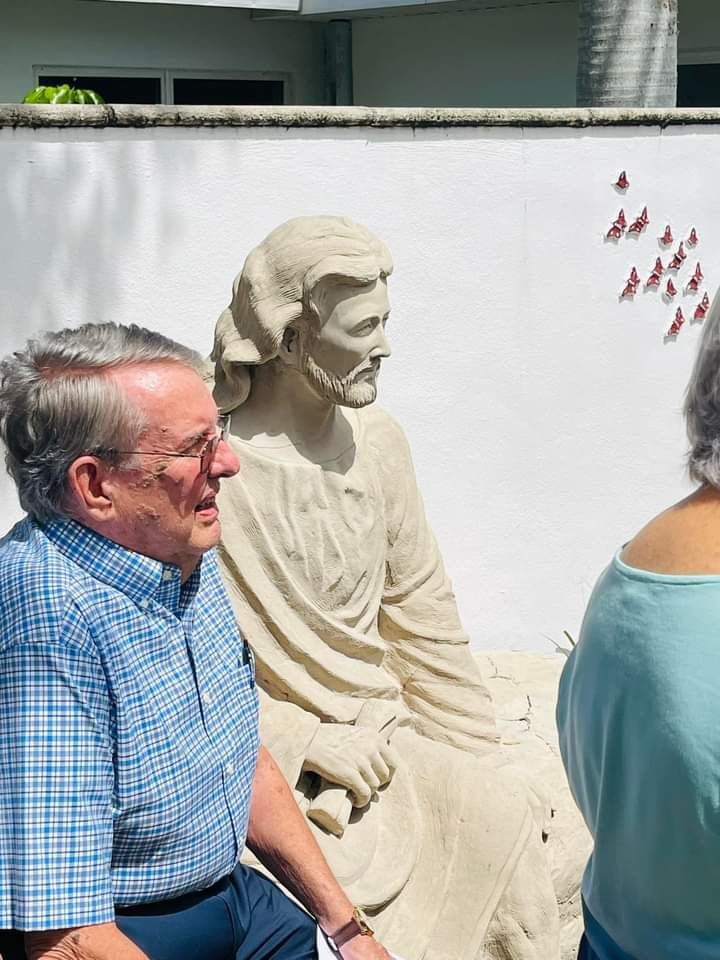 Man with glasses beside a seated Jesus statue against white wall with decorative red birds.