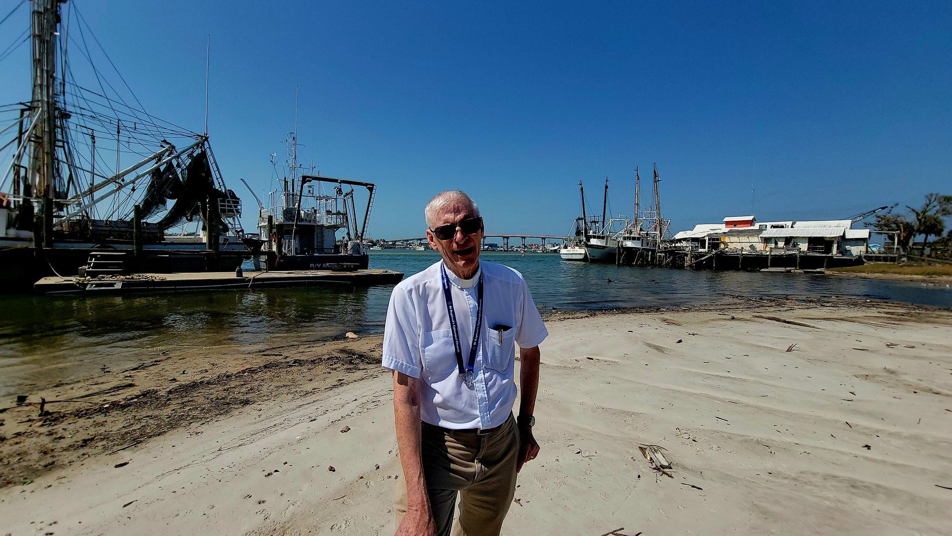 Man in white shirt stands on sandy shore with boats and blue sky.