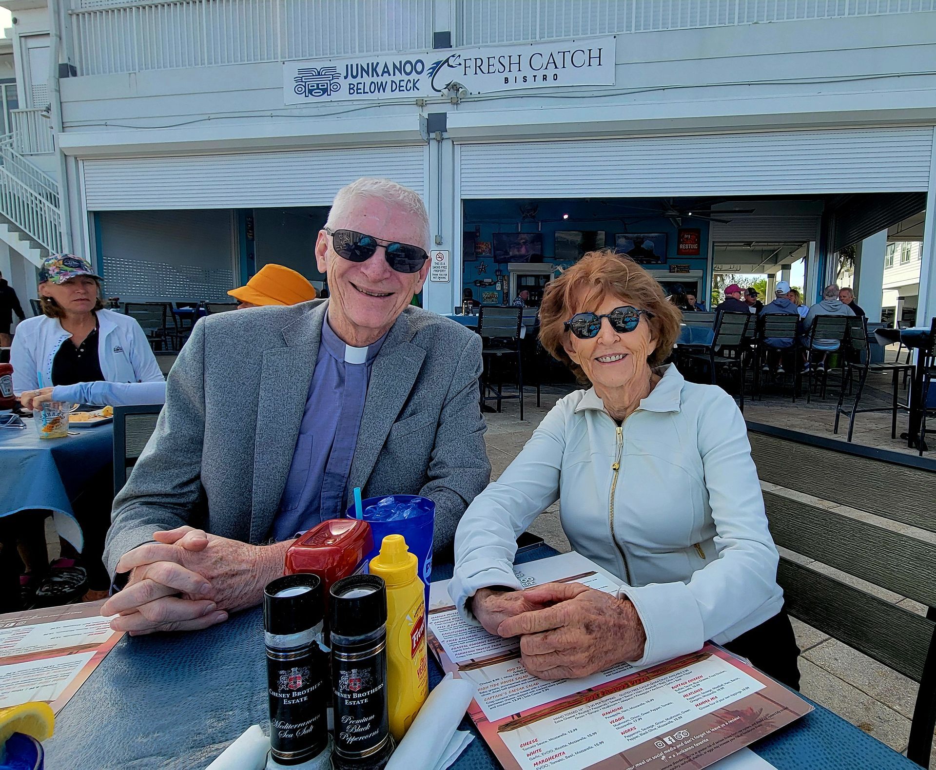 A smiling couple at an outdoor restaurant, man wearing a collar and sunglasses, woman in a jacket.