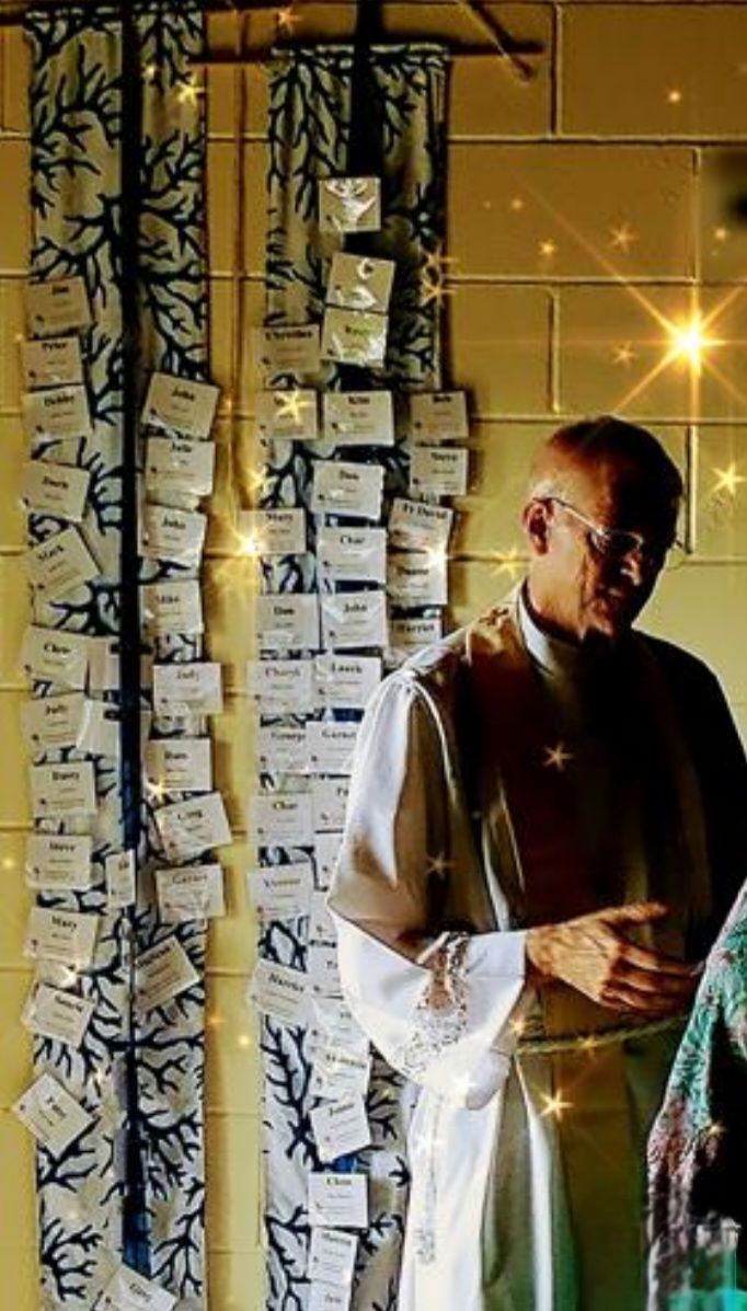 Man in white robe stands by wall with name cards hanging on decorative fabric.