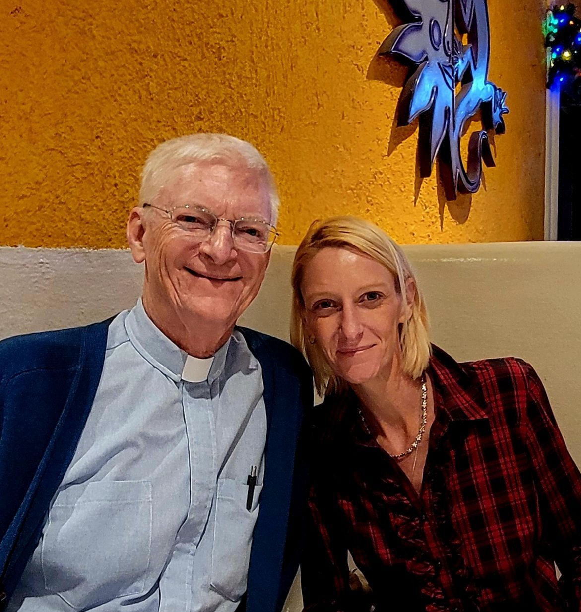 An older man in a clerical collar and a blonde woman smile together, indoors with a decorative wall.