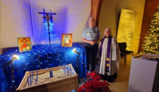 Two people by a religious display, a cross, candles, and a Christmas tree. One wears a clerical robe.