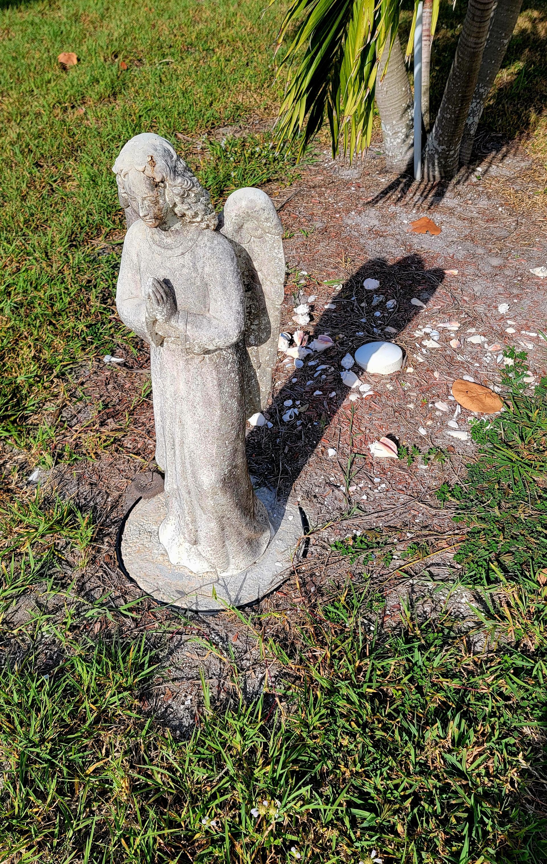 A weathered stone angel statue stands in a grassy yard, with a shadow cast to the side.