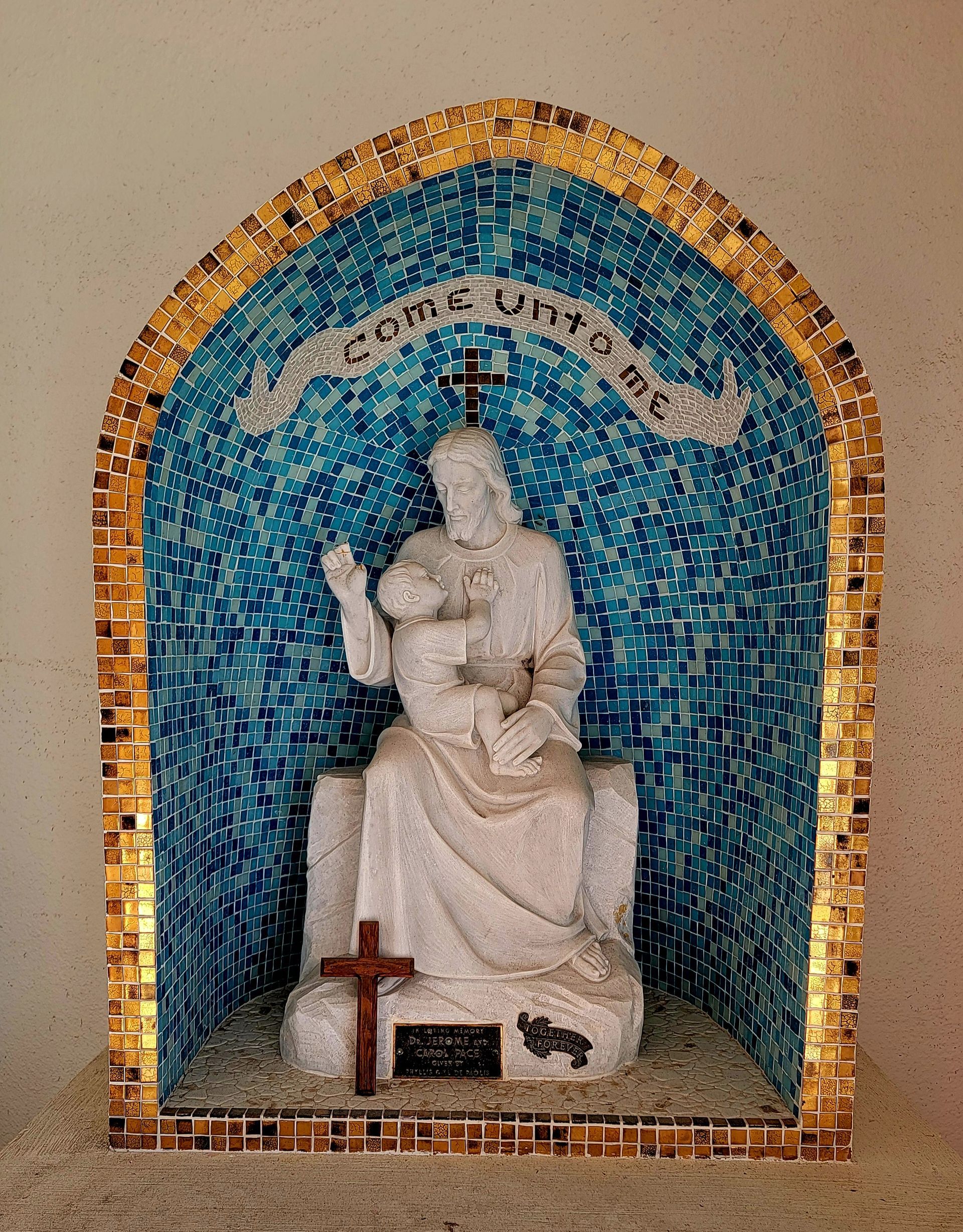 Statue of Jesus holding a child within a blue mosaic archway, with a cross below.