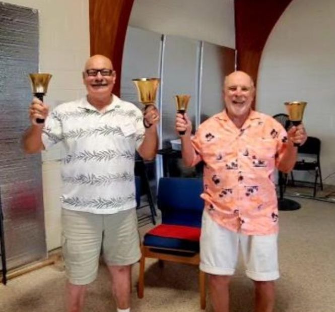 Two men holding handbells, smiling, in a room. One in white, one in pink shirt.