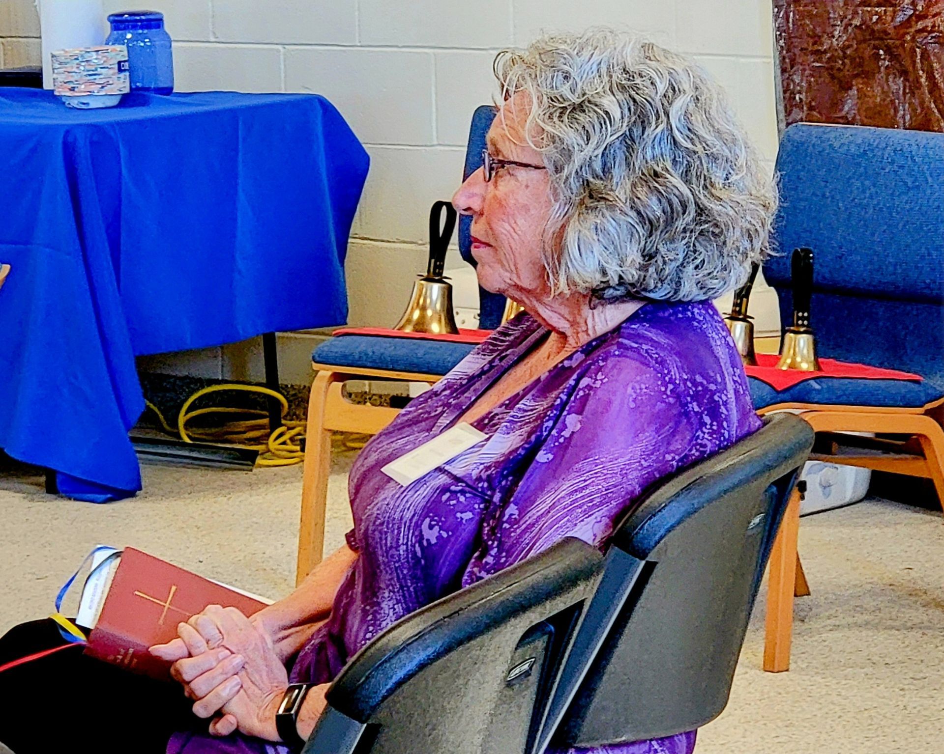 Woman with gray curly hair in a purple shirt seated, holding a book, near handbells and blue tables.
