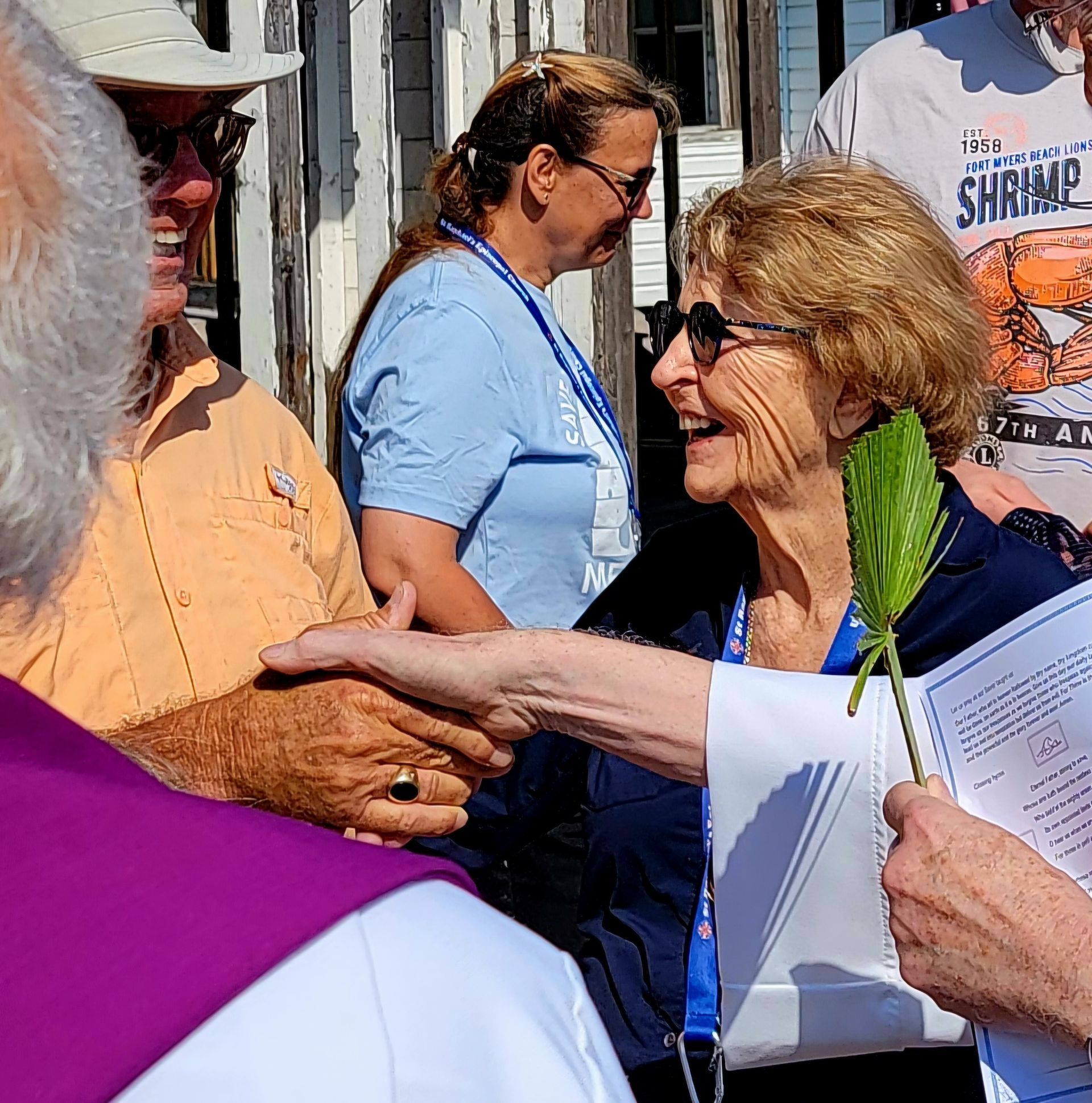 People greeting each other outdoors, smiling. Woman in sunglasses holds a green leaf.