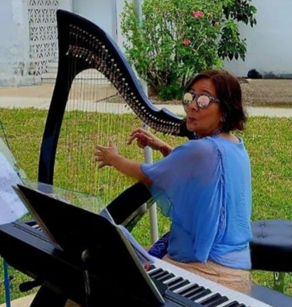 Woman playing a large black harp outdoors, wearing blue shirt and sunglasses. Piano and music sheets in front.