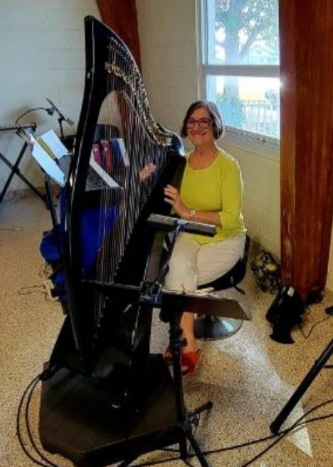 Woman smiling, seated, playing a large black harp indoors by a window.