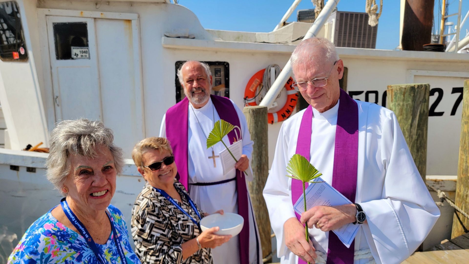 Two women and two clergy, one holding a bowl, on a dock near a white boat.